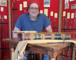 An older white man sits behind a wooden table. He's wearing a red hat, glasses, jeans, and a blue t-shirt. On the table is a larger leather-bound ledger, and atop the ledger is a bamboo rod. A display behind him features more bamboo rods against a red backdrop, along with illustrations and text panels.