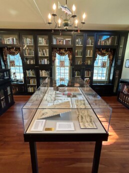 Looking at the narrow end of a long display case with a plexiglass cover. The case contains maps and placards with text on them. The case is in a library, and a chandelier decorated with a trout and flies sits overheard.
