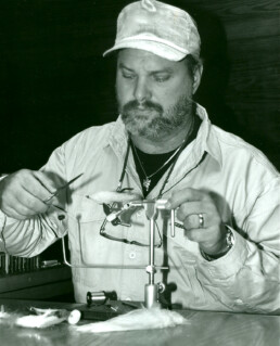 A black and white photo of a bearded man tying a streamer fly.