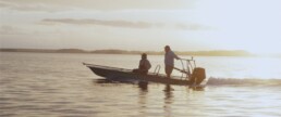 Two men, backlit by the sun, take a skiff across a body of water.
