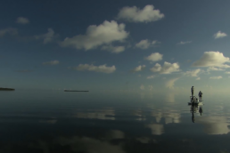 A large expanse of water reflects the sky and clouds. On the right side two people stand on a skiff, one on the deck and one on the poling platform.